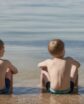 two boys sitting on the shore of a body of water