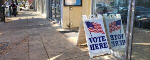 sidewalk with a sandwich board sign that says "vote here" with an american flag. the text is mirrored in a window.