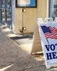 sidewalk with a sandwich board sign that says "vote here" with an american flag. the text is mirrored in a window.