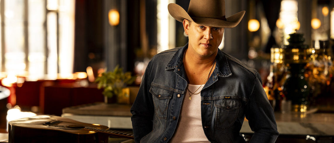 man wearing a cowboy hat, jean jacket and a shirt standing in front of a guitar on a table