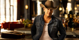 man wearing a cowboy hat, jean jacket and a shirt standing in front of a guitar on a table