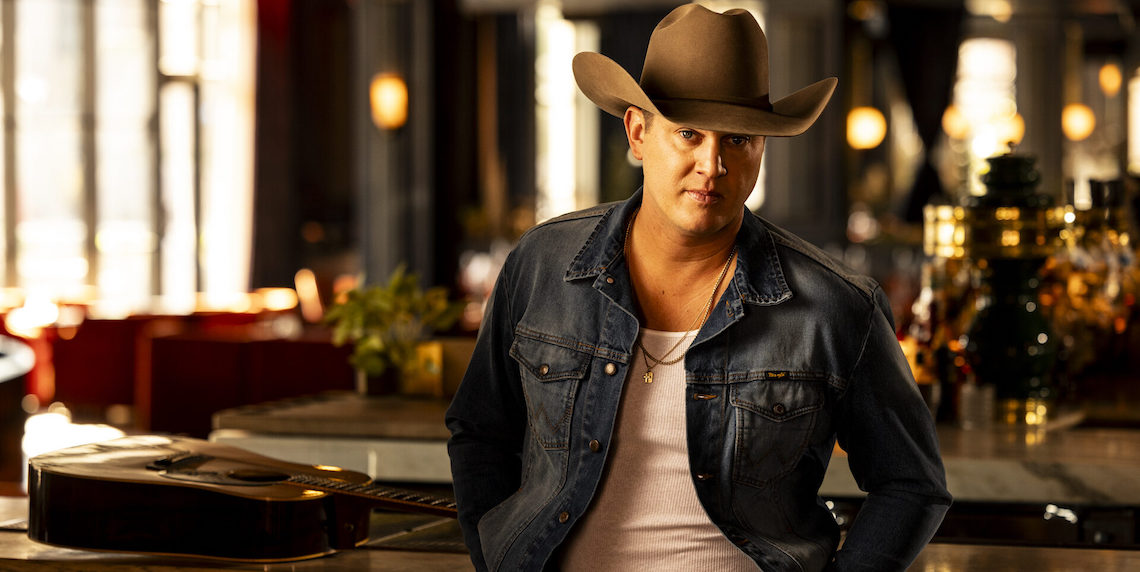 man wearing a cowboy hat, jean jacket and a shirt standing in front of a guitar on a table