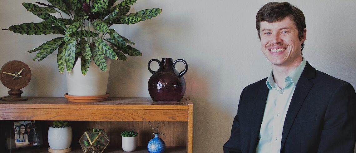 man smiling as he sits next to a book shelf