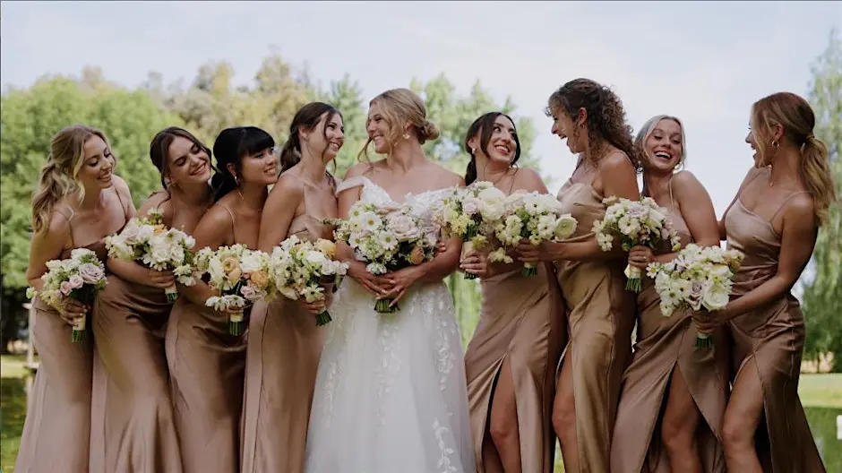 A bride in a white dress surrounded by eight bridesmaids in brown gowns, holding bouquets.