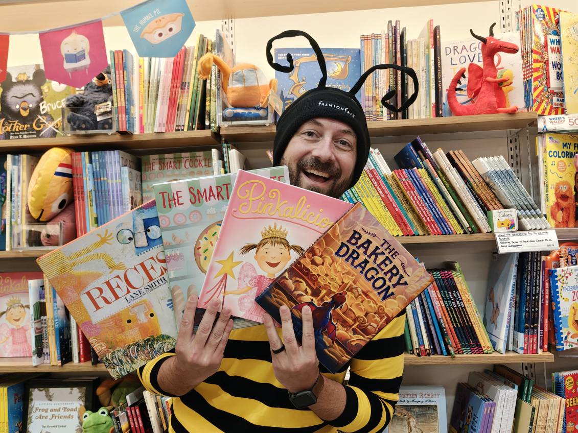 Mr.B is dressed in a bumble bee costume with four children's books in his hands. He is sitting in front of a book shelf filled with more children's books.