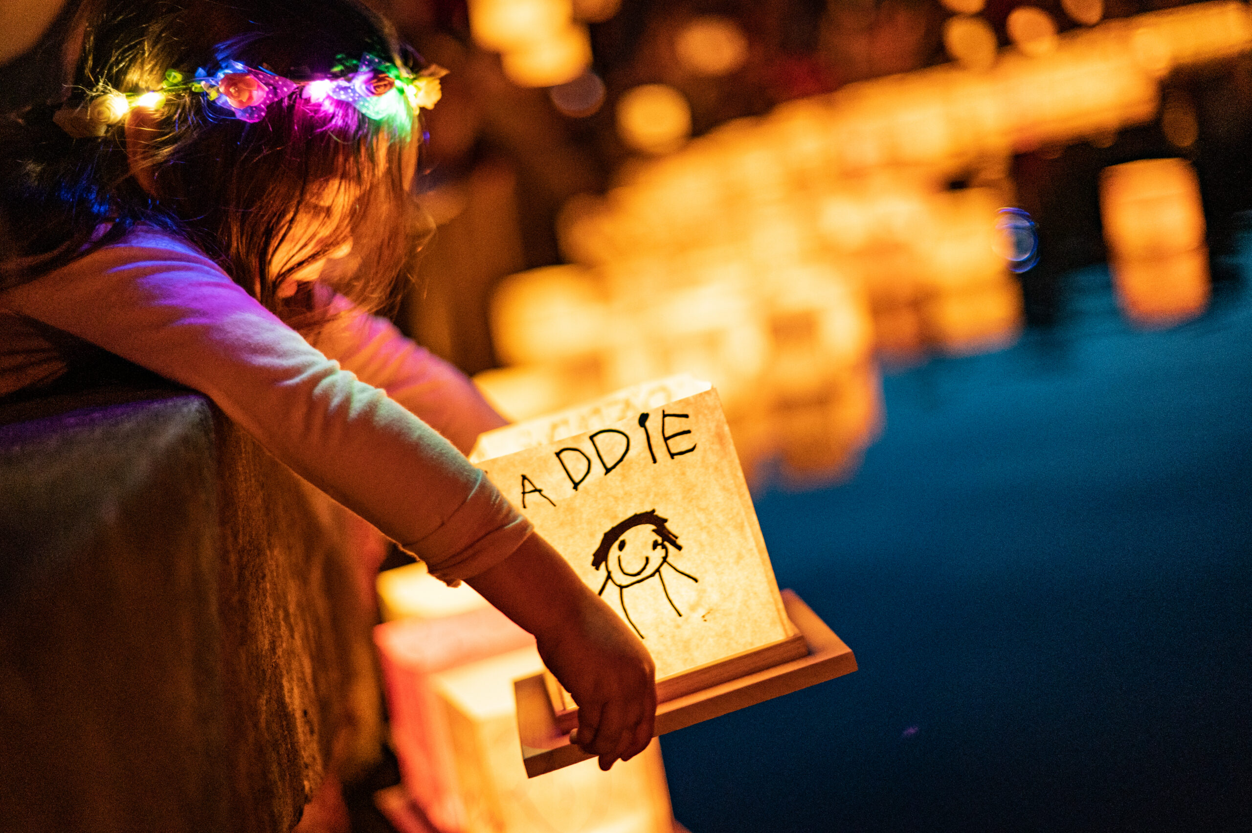 A photo of a little girl in an illuminated flower-crown getting ready to put a paper lantern with the name Addie written on it into the water.