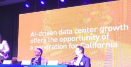 a woman speaks at a podium onstage with two men looking on with a slide deck behind them