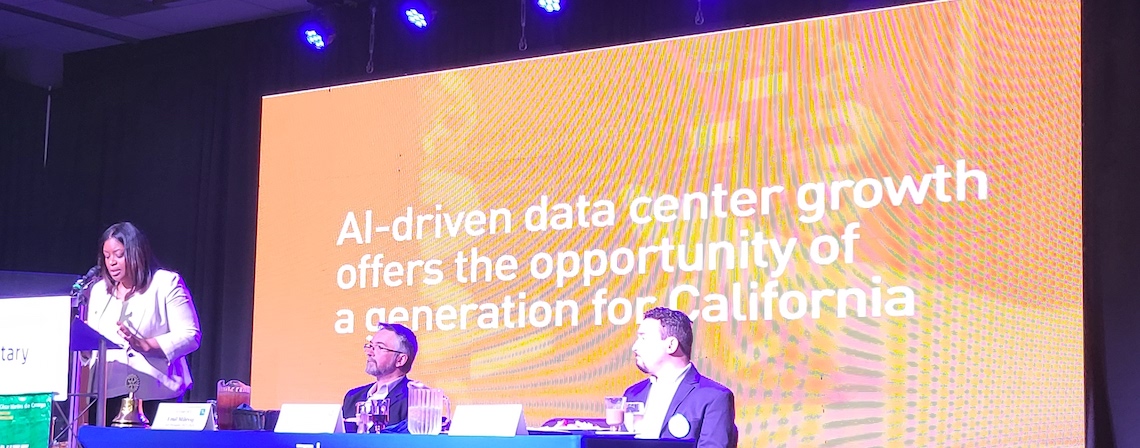 a woman speaks at a podium onstage with two men looking on with a slide deck behind them