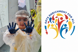 Student wearing protective goggles and a smock holds up paint-covered hands during an Arts Enrichment for All art activity.