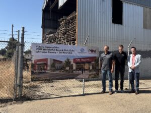 Three men stand beside a chain-link fence in front of a banner showing a rendering of the future Del Rey Boys & Girls Club.