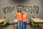 A man and a woman in orange shirts stand smiling in front of a hand-drawn "Raw Fresno" mural inside the restaurant, with wooden tables and green chairs on either side.