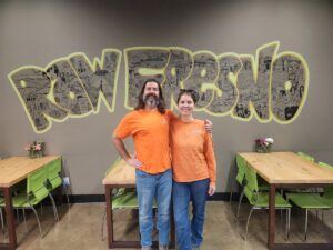 A man and a woman in orange shirts stand smiling in front of a hand-drawn "Raw Fresno" mural inside the restaurant, with wooden tables and green chairs on either side.