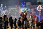 Visitors walk past a Halloween yard display with colored lights, artificial fog and people dressed as horror movie characters.