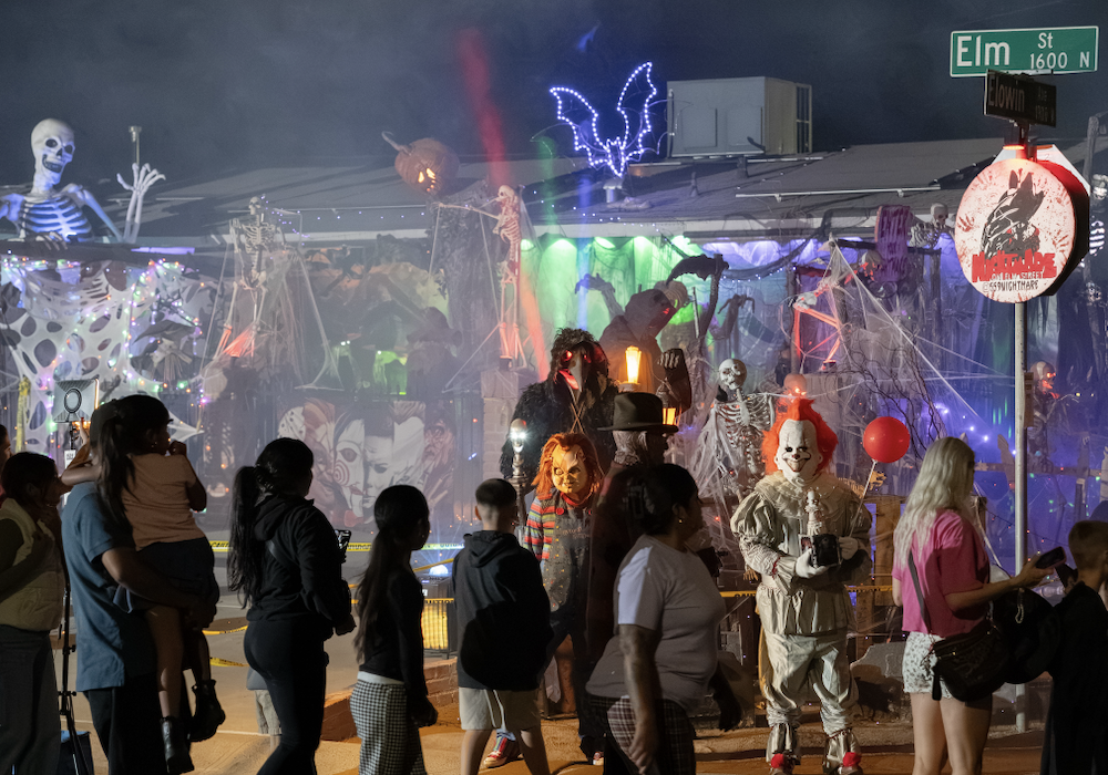 Visitors walk past a Halloween yard display with colored lights, artificial fog and people dressed as horror movie characters.
